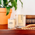 Two candles from Ninth House Studio on a wooden surface with a plant and books in the background.