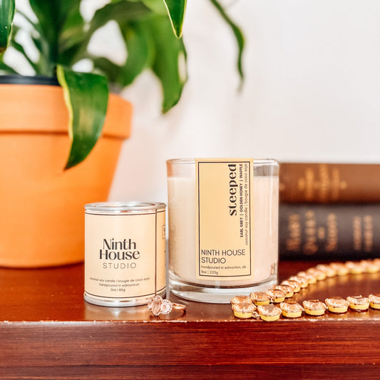 Two candles from Ninth House Studio on a wooden surface with a plant and books in the background.
