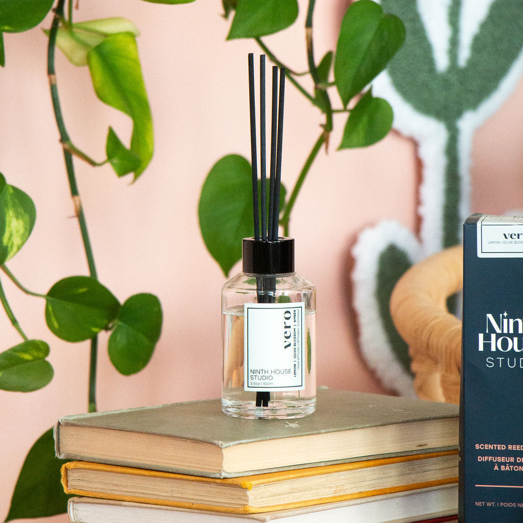 A Vero Reed Diffuser bottle with black reeds sits on a stack of books on a wooden table, with a Vero Reed Diffuser box next to it and a pink wall with green plant leaves in the background.