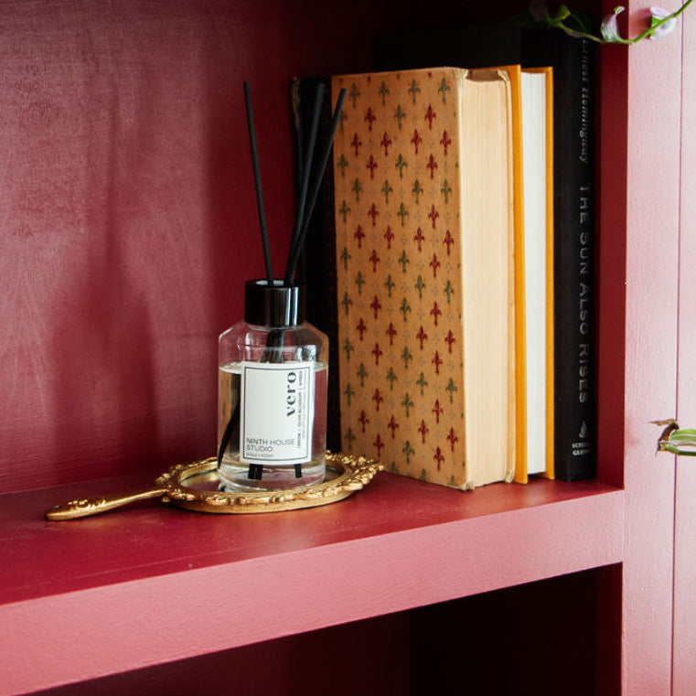 A Vero reed diffuser bottle with a clear glass body, black reeds, and a light green label reading 'Vero' sits on a small gold tray on a pink shelf next to patterned books.