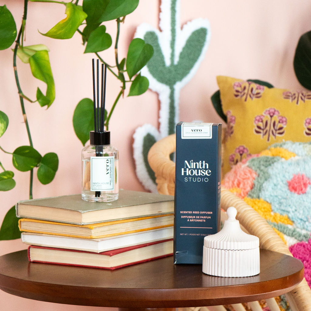 A reed diffuser bottle with black reeds sits on a stack of books on a wooden side table, next to a dark blue Ninth House Studio product box and a white ribbed container, in a styled room with a pink wall, green cactus wall hanging, and patterned cushions.