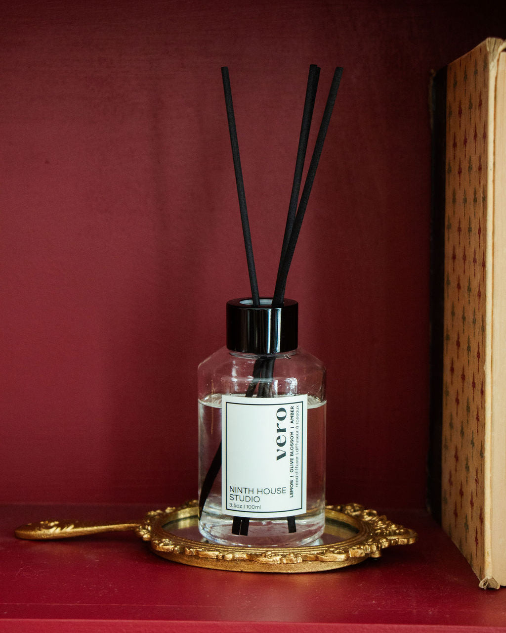 A clear glass reed diffuser bottle with a black cap and four black reeds, featuring a white label that reads 'Vero' and 'Ninth House Studio', displayed on an ornate gold tray against a dark red background.